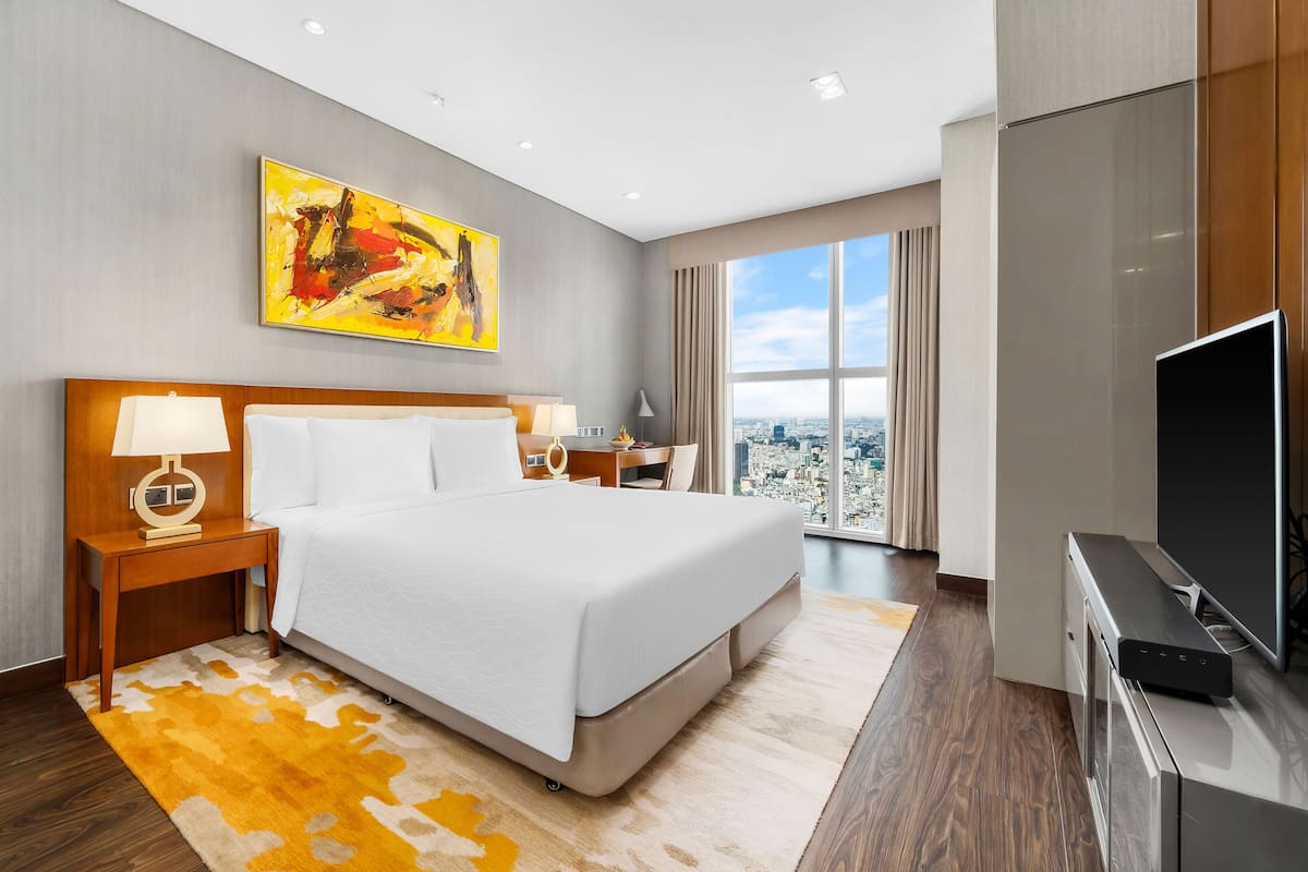 A modern hotel room with a large bed, a flat-screen TV, a desk with a lamp, and a city view through the window.