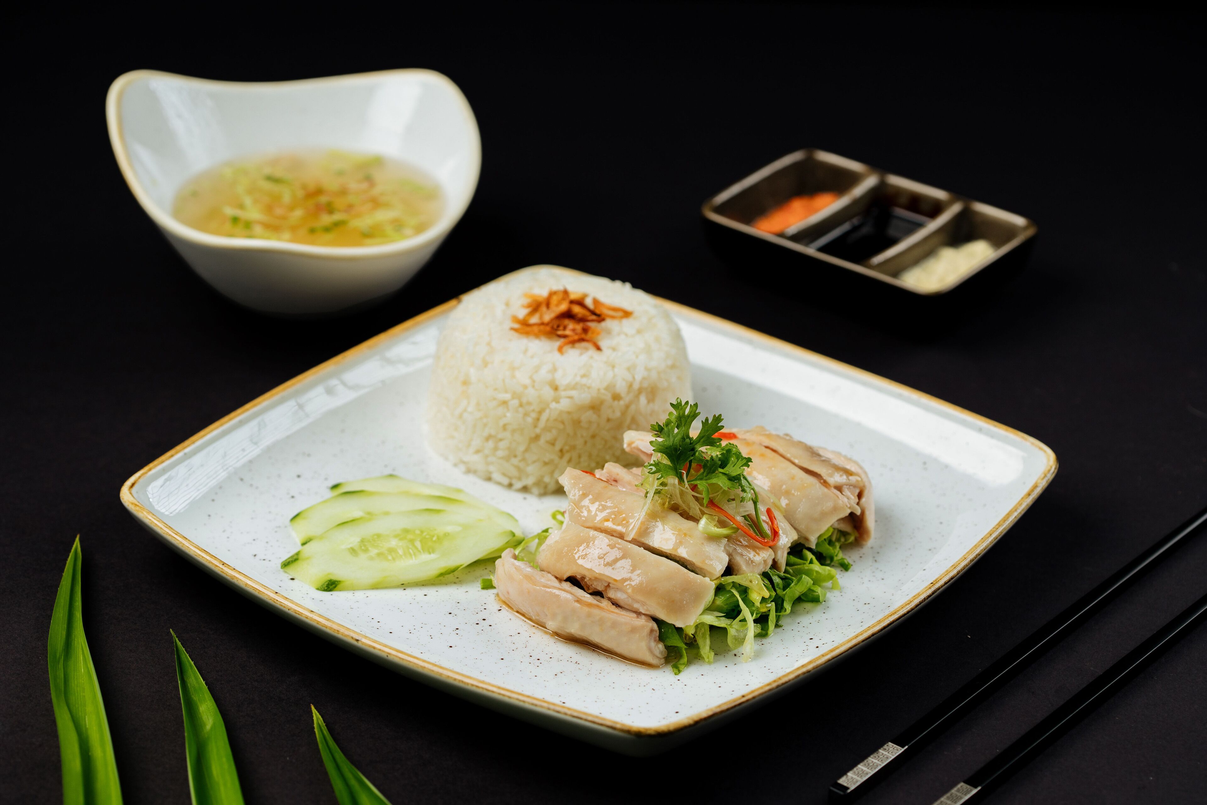 A plate of food with rice, sliced chicken, cucumber, and lettuce, accompanied by a small bowl of soup and a dish of sauces.
