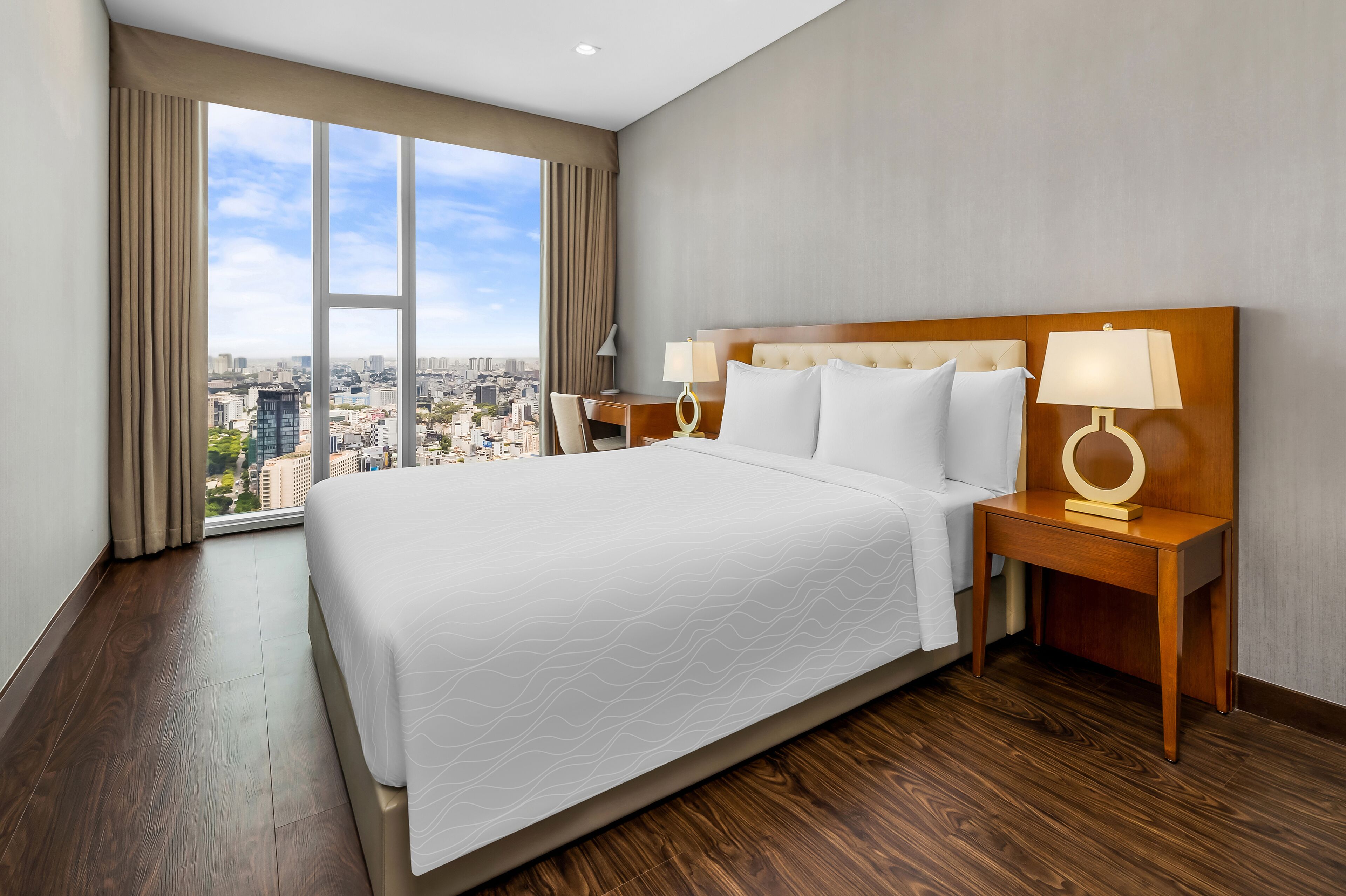 A hotel room with a large bed, two bedside tables with lamps, a view of the city through large windows, and a cityscape visible outside.