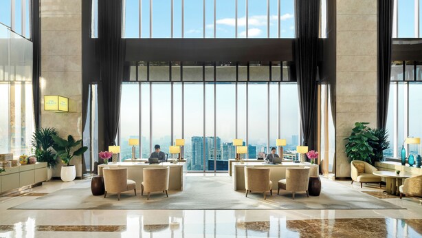 A spacious lobby with a high ceiling, large windows, and a view of the cityscape. There are several seating areas and a reception desk with two staff members.