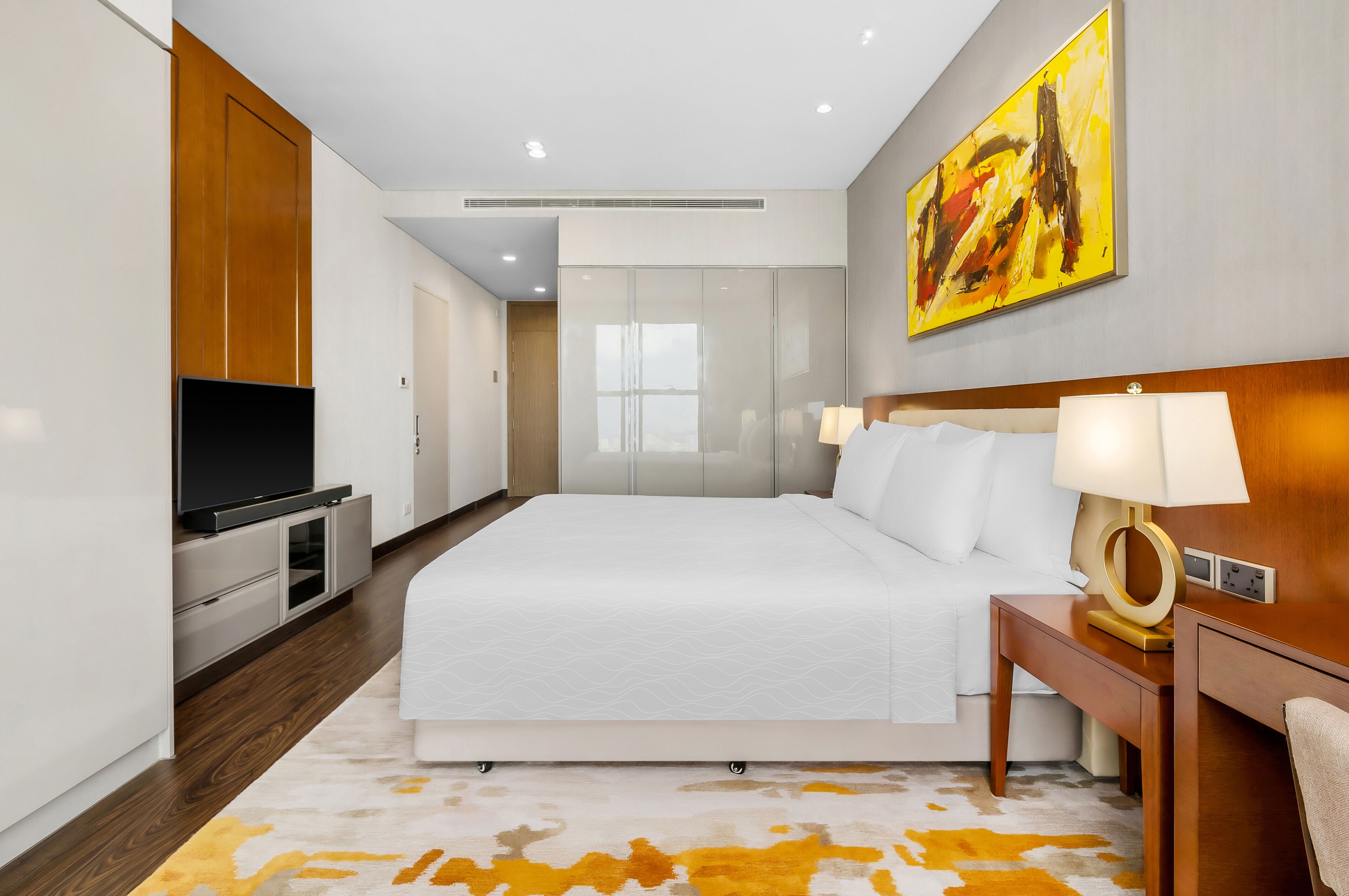 A modern hotel room with a large bed, a flat-screen TV, a desk with a lamp, and a painting on the wall.