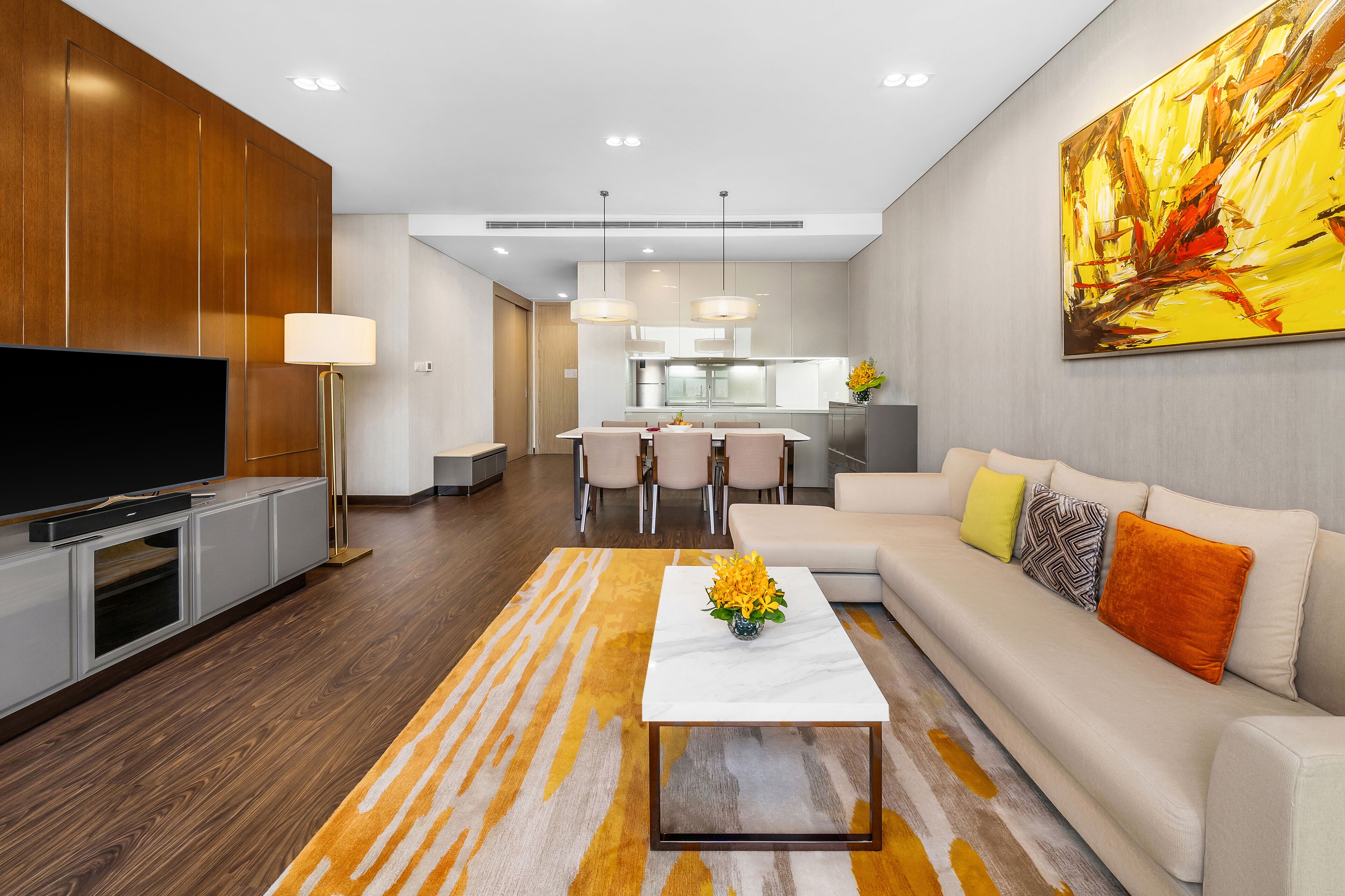 A modern living room with a flat-screen TV, a coffee table with a vase of flowers, a sofa with cushions, and a dining area in the background.