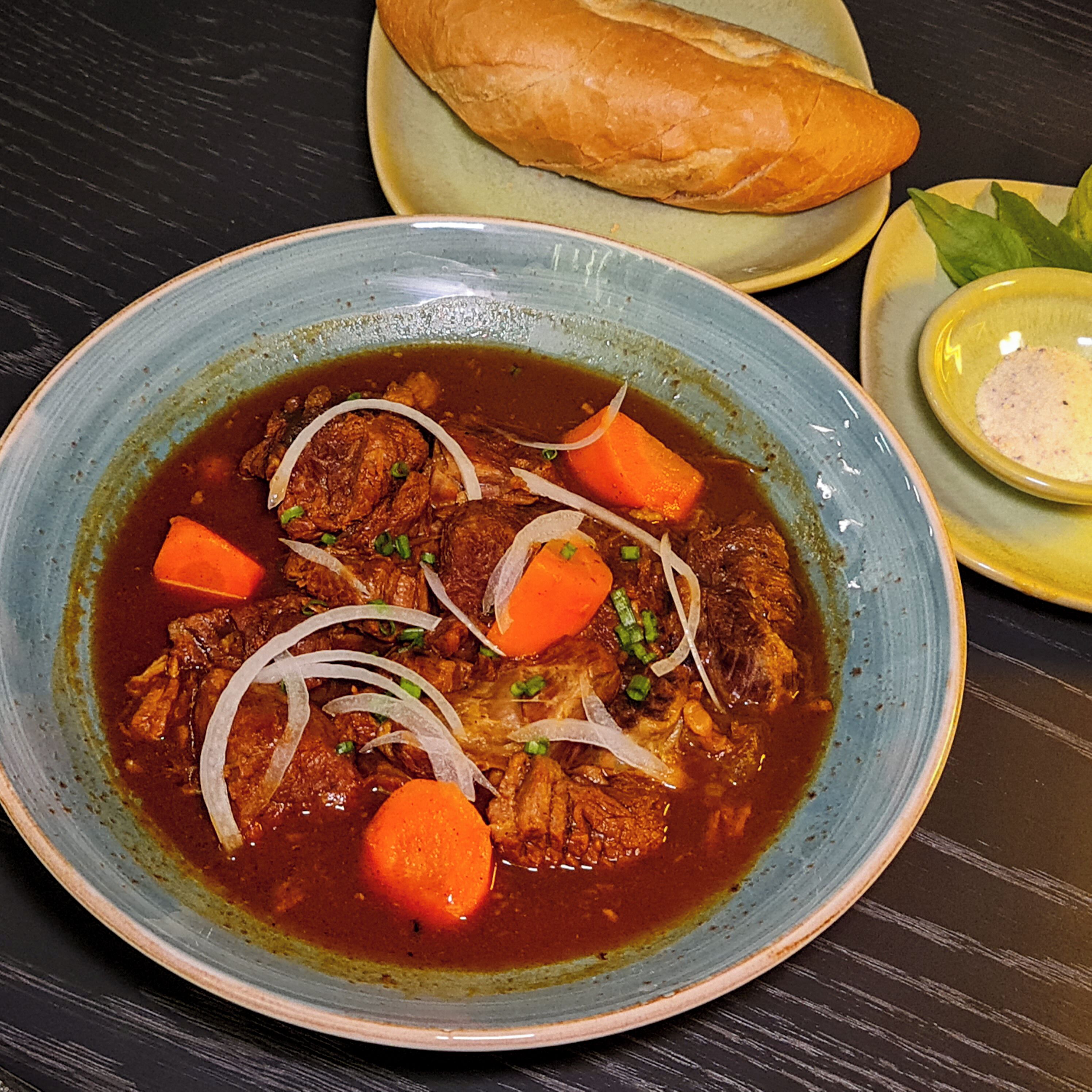Un plato de guiso con carne, zanahorias y cebollas, acompañado de una baguette y un recipiente pequeño de salsa sobre una mesa de madera oscura.