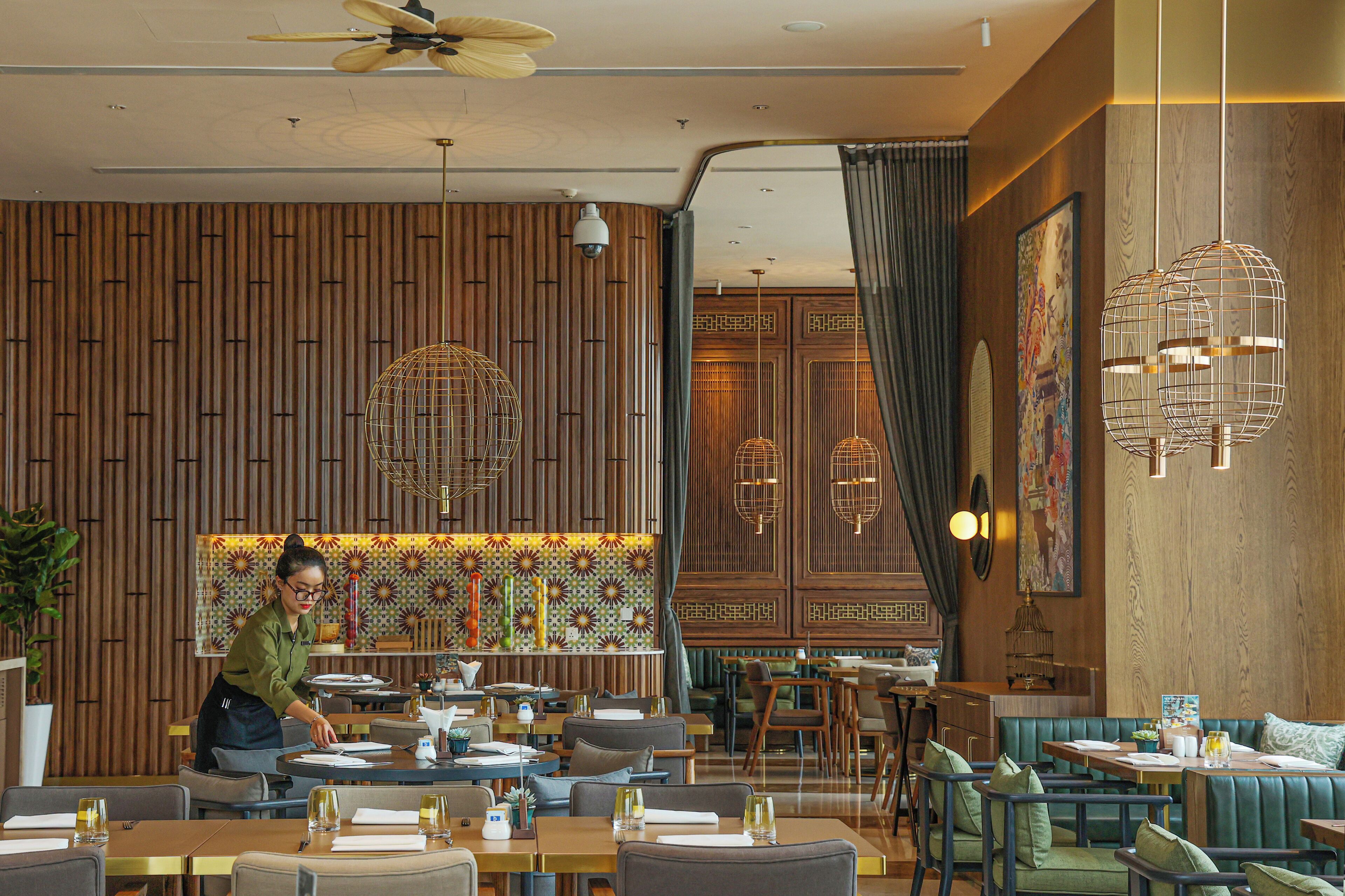 Interior de restaurante con paredes de paneles de madera, asientos tapizados en verde y personal de servicio preparando mesas.