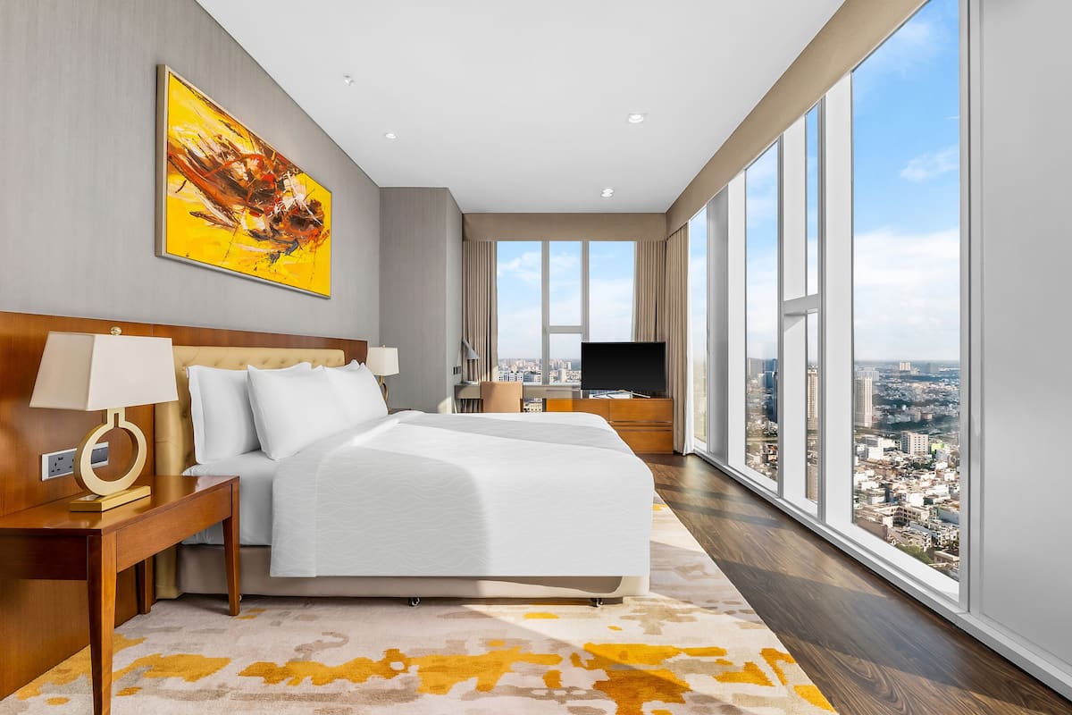 A modern hotel room with a large bed, a TV, and a city view through large windows.
