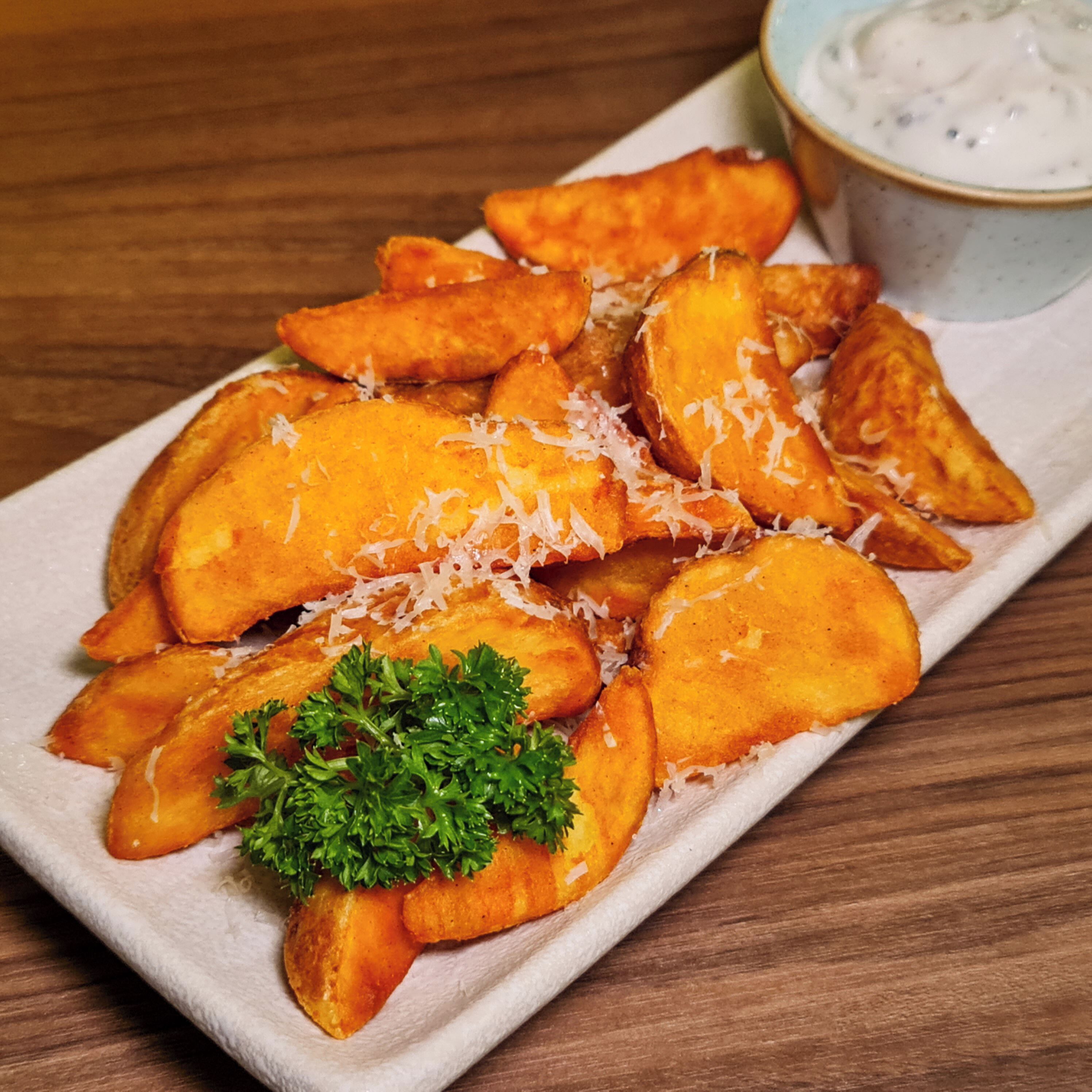 A plate of golden-brown potato wedges garnished with shredded cheese and parsley, accompanied by a small bowl of dipping sauce.