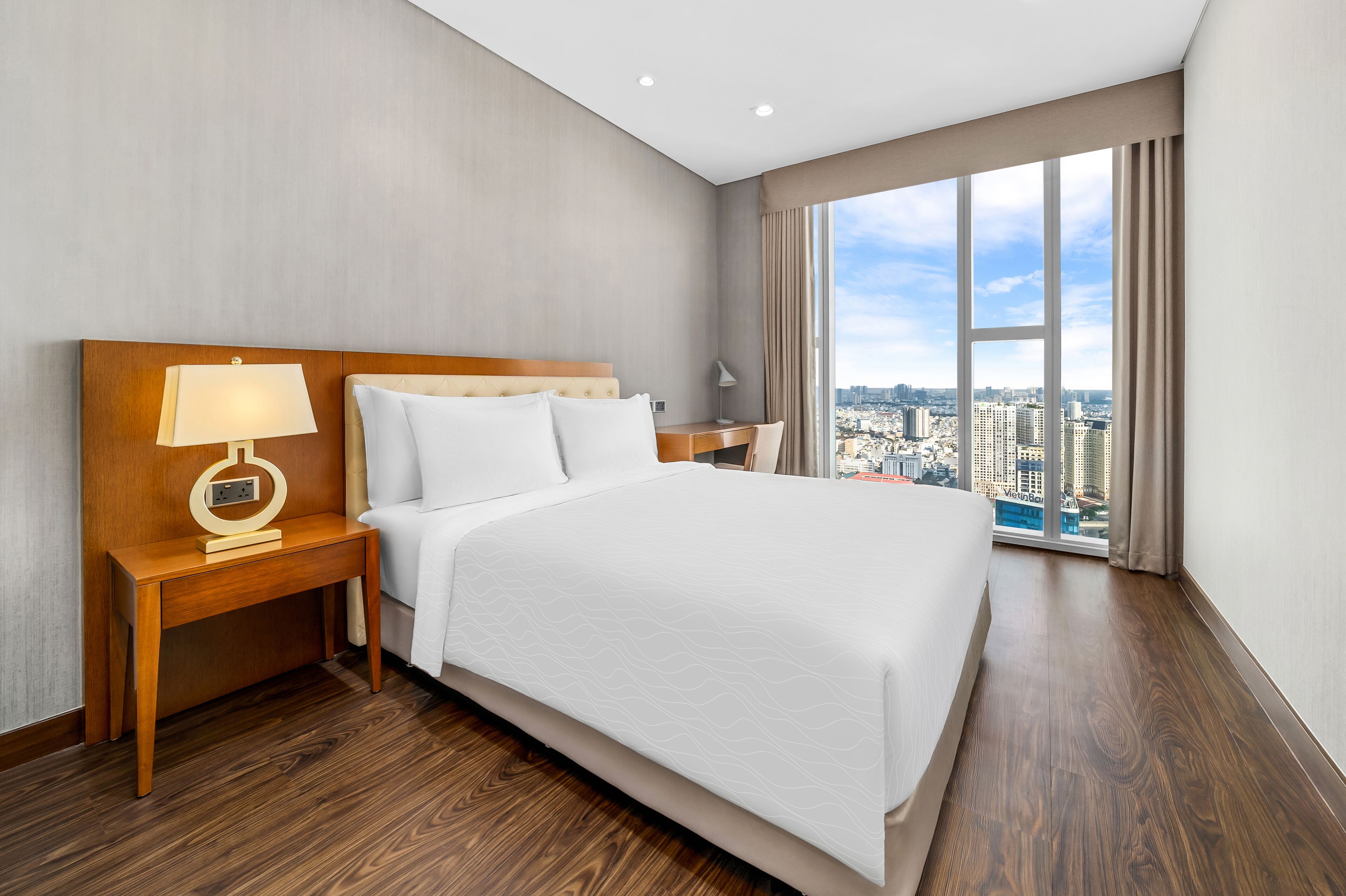 A hotel room with a large bed, a wooden nightstand with a lamp, a desk, and a view of a cityscape through large windows.