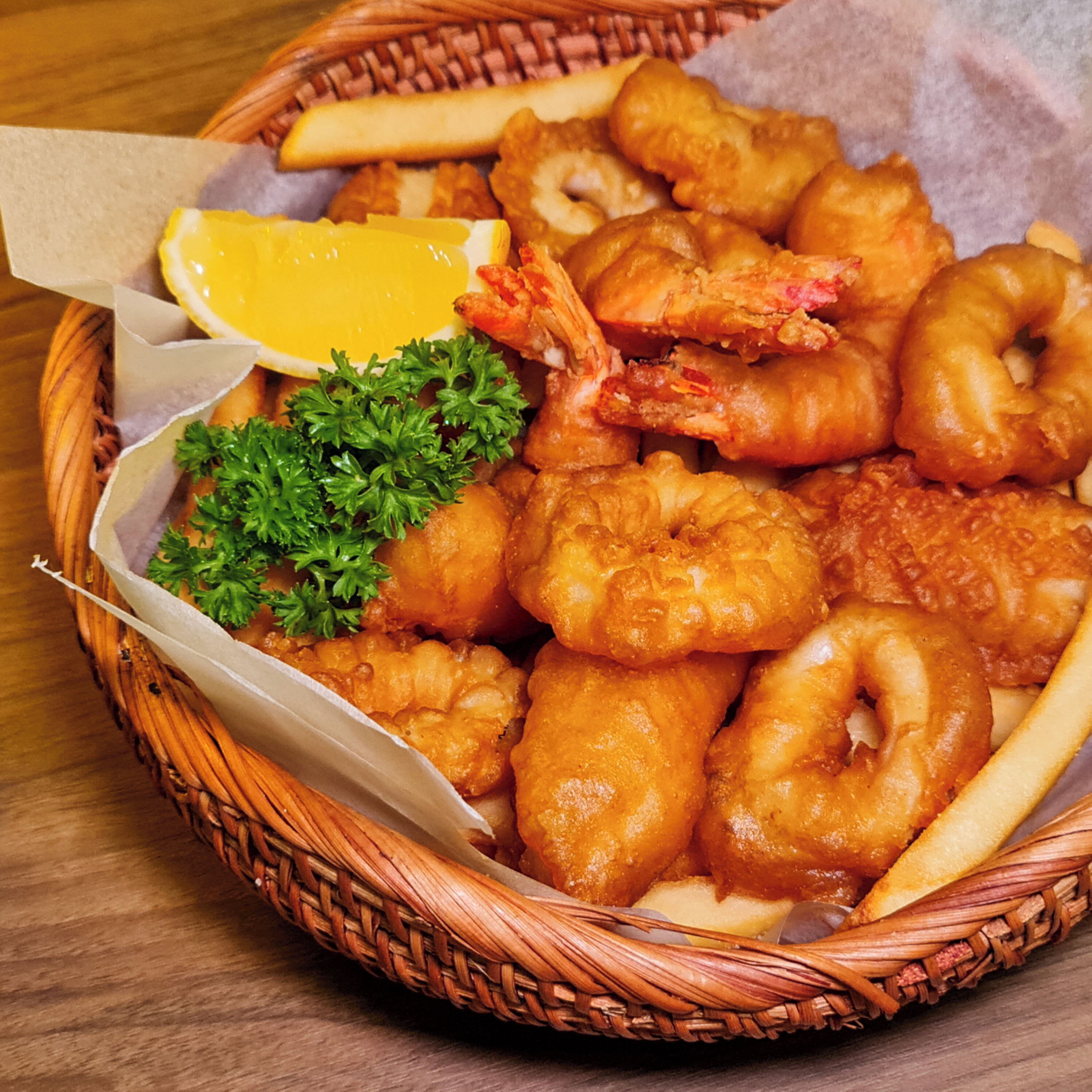 Un panier de fruits de mer frits, comprenant des crevettes, des coquilles Saint-Jacques et des rondelles de calamars, servi avec des quartiers de citron et du persil.