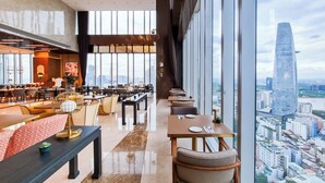 A modern hotel lobby with a dining area, large windows offering a city view, and a staircase leading to an upper floor.