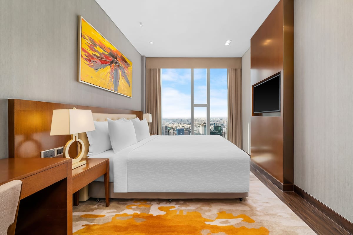 A hotel room with a large bed, a desk with a lamp, a TV, and a city view.