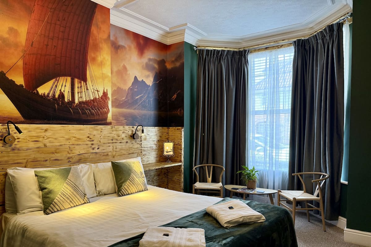 Quarto de hotel com uma cama grande, cabeceira com mural, pequena mesa com planta e janela com cortinas.