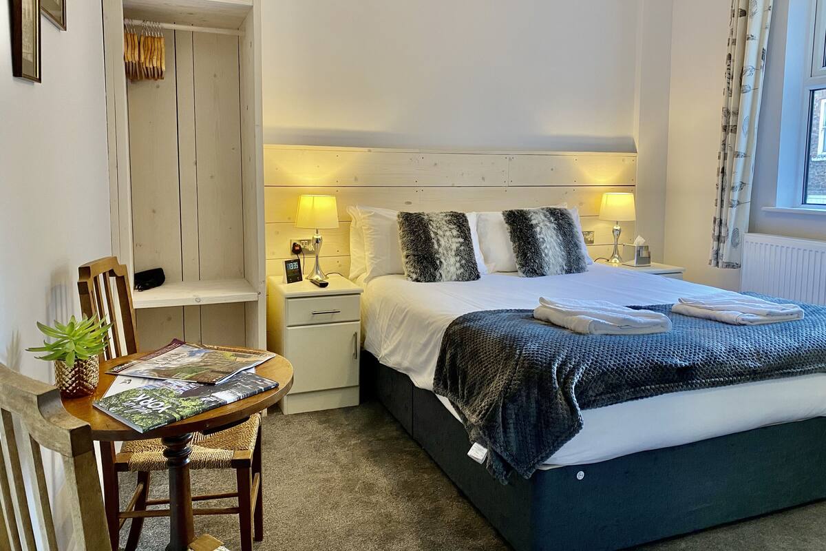 Quarto com cama, mesas de cabeceira, um banquinho, uma cadeira, uma mesinha com uma planta e uma janela com cortinas.