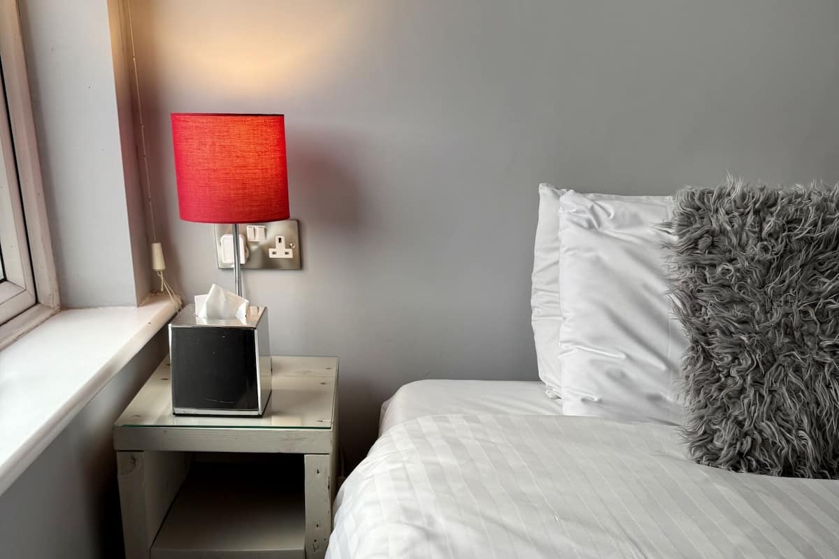 Quarto de hotel com cama, mesa de cabeceira, luminária, caixa de lenços e janela.