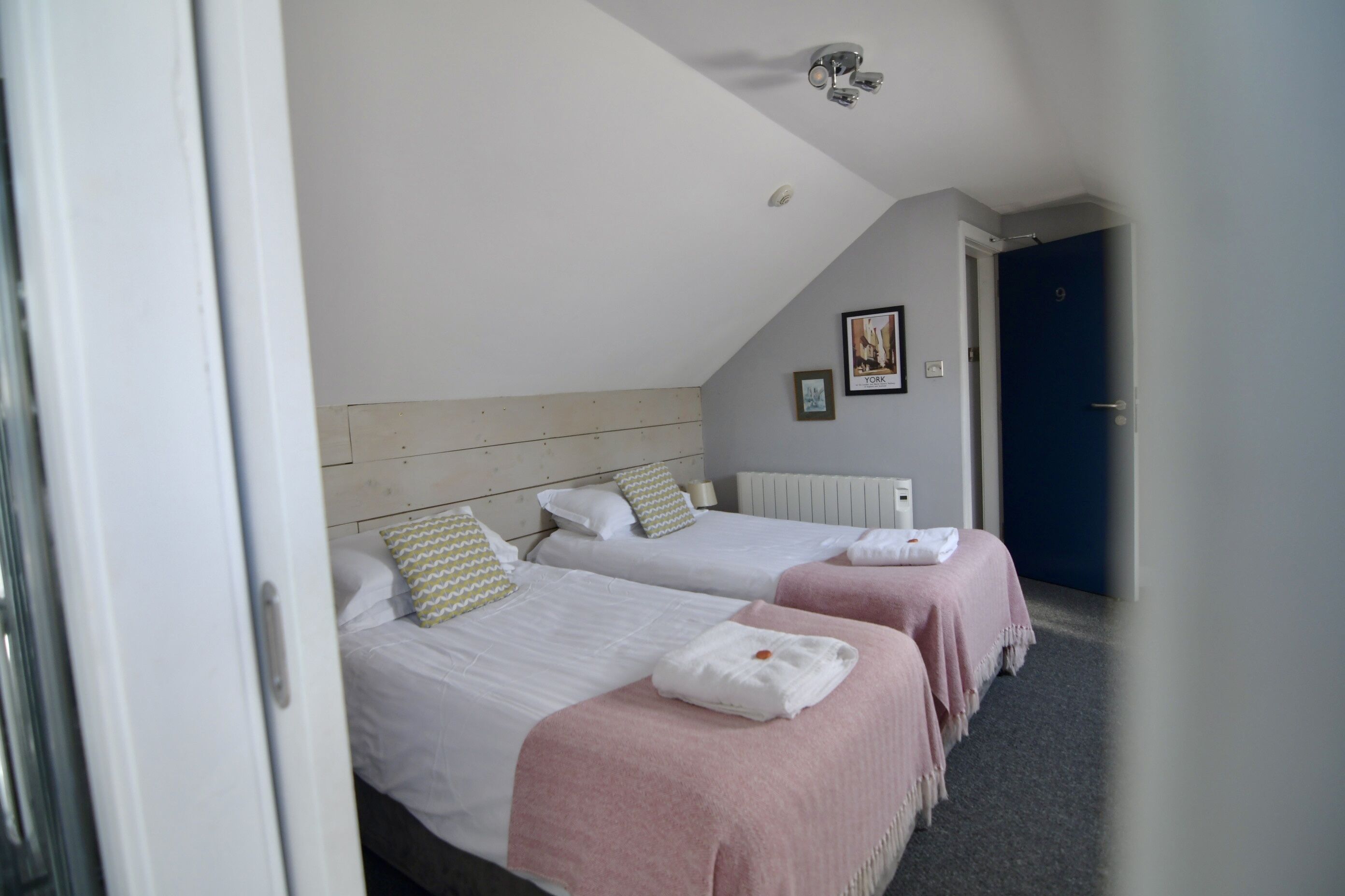 Une chambre avec deux lits, un plafond blanc, une porte bleue et un tableau encadré au mur.