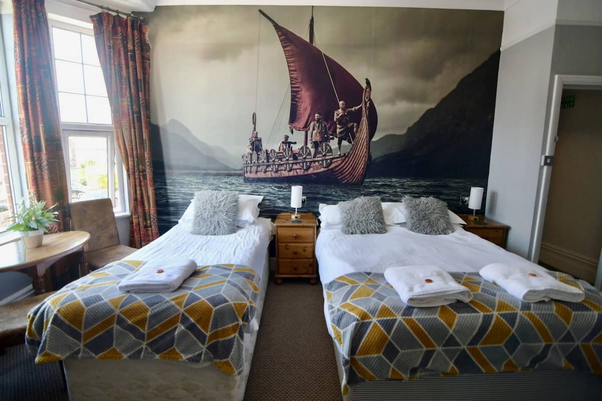 Quarto de hotel com duas camas, um mural de um navio a vela, uma escrivaninha e uma janela com cortinas.