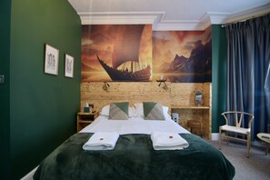 Ett hotellrum med en stor säng, ett träfärgat sänggavel, gröna väggar och en väggmålning som föreställer en segelbåt.
