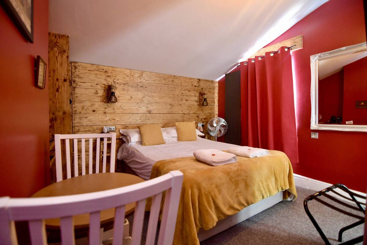 Quarto com cabeceira de madeira, cama com cobertor amarelo, cadeira branca e cortina vermelha.