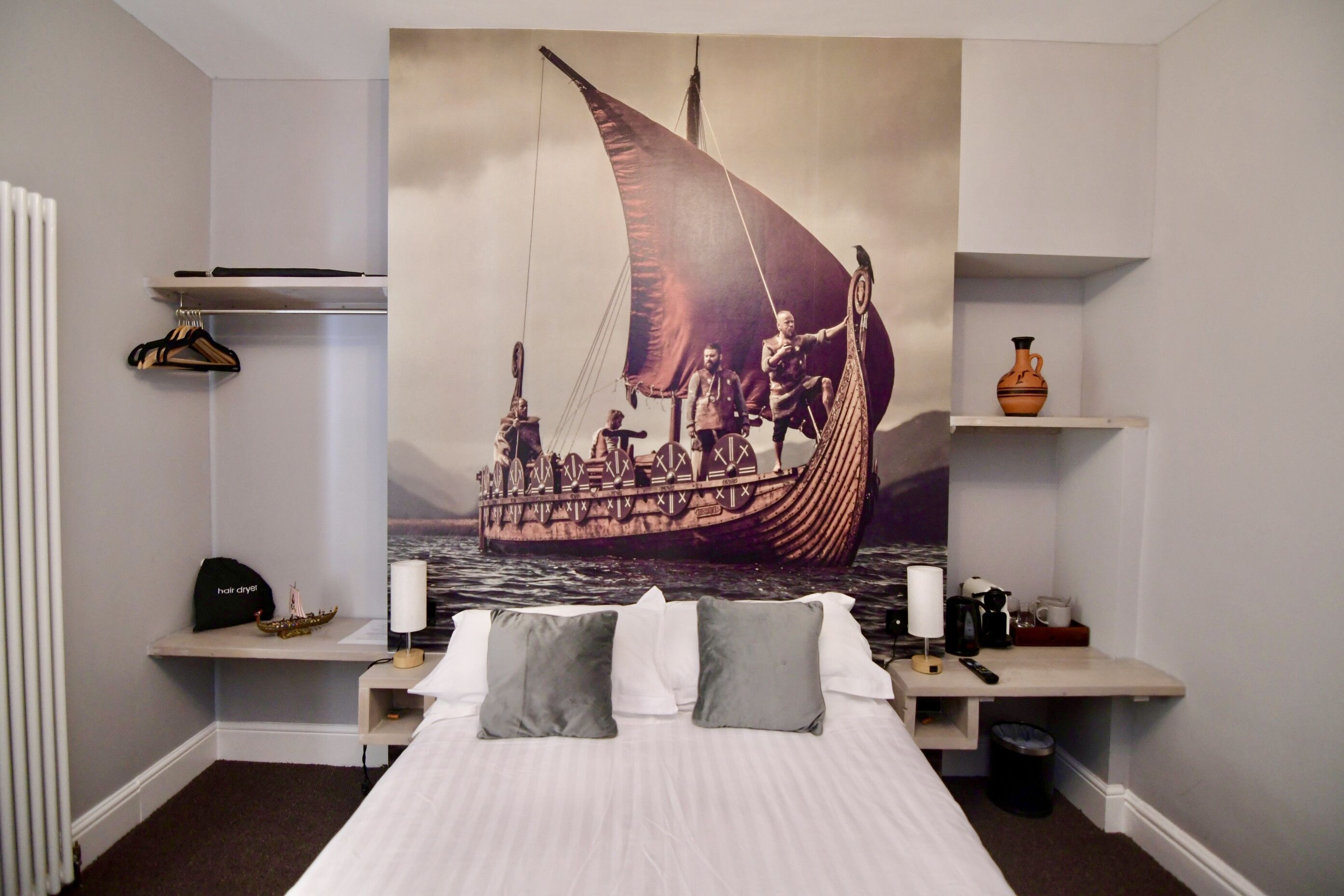Un dormitorio con una cama, almohadas, una mesita de noche, un estante en la pared con un florero, y un cuadro grande de un barco.