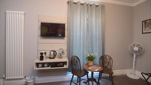 A room with a TV mounted on the wall, a radiator, a fan, two wooden chairs, a small table with a plant, and a picture frame on the wall. - Vikings Accommodation (York)