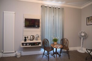 Quarto com televisão na parede, radiador, ventoinha, duas cadeiras de madeira, uma mesa pequena com uma planta e um porta-retrato na parede.