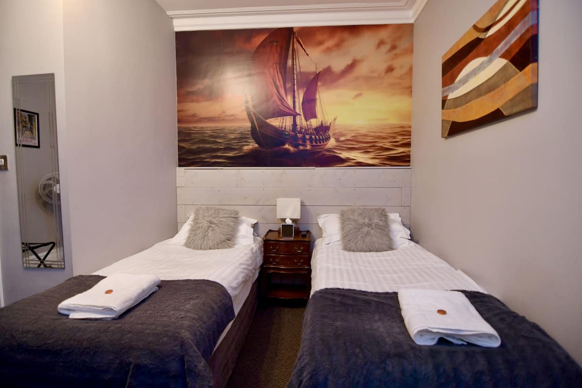 Quarto de hotel com duas camas, um quadro de um veleiro na parede e uma pequena mesa de cabeceira de madeira.