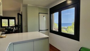 Une cuisine avec vue sur la mer, un réfrigérateur et une fenêtre équipée de stores.