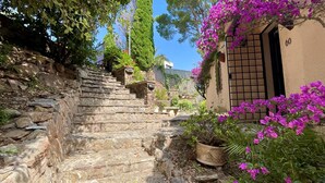 Un escalier en pierre mène à un bâtiment doté d’une porte, le tout étant entouré d’une végétation luxuriante et de fleurs violettes.