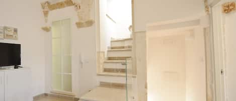 A spacious room with a staircase leading to an upper floor, a flat-screen TV on a white cabinet, and a glass door.
