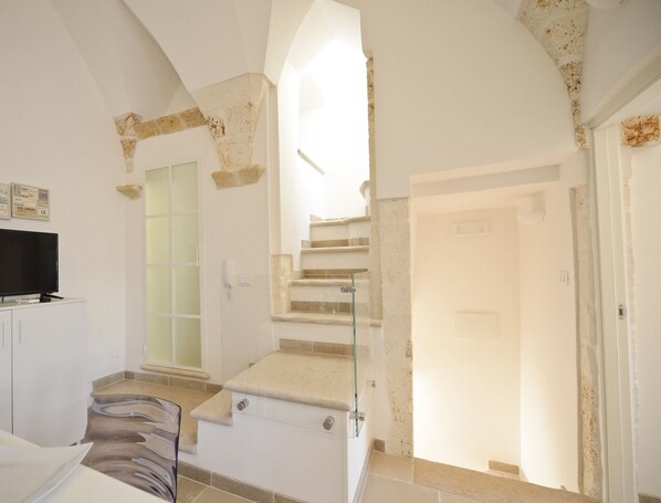 A spacious room with a staircase leading to an upper floor, a flat-screen TV on a white cabinet, and a glass door.
