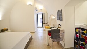 A hotel room with a bed, a desk with a chair, a small table with fruits, a mirror, and a colorful shower curtain. - Scirocco Apartment with Terrace by Wonderful Italy (Ostuni)