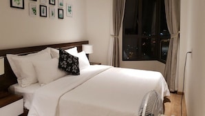 Un lit bien fait, avec du linge de lit blanc, une tête de lit en bois et une table de chevet avec une lampe. Des reproductions botaniques encadrées ornent le mur, et une fenêtre avec des rideaux offre une vue sur la ville la nuit.