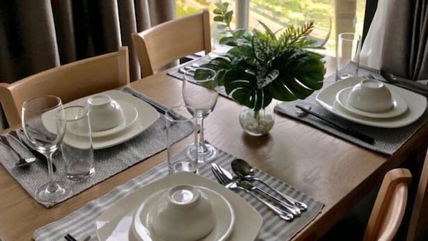 Une table à manger dressée pour deux personnes, avec assiettes, couverts et verres, offrant une vue pittoresque sur un plan d’eau et la verdure environnante.