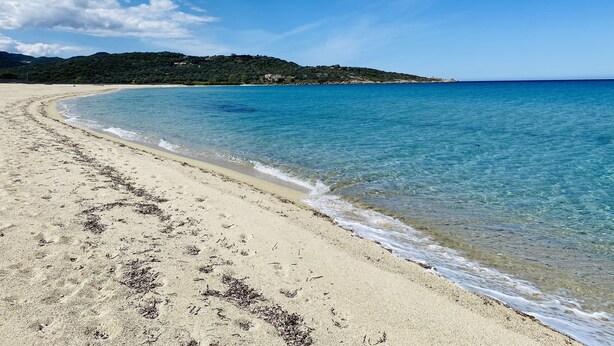 Una playa con agua azul cristalina, orilla arenosa y un paisaje montañoso al fondo.