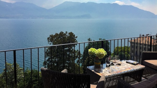 Un balcon avec vue sur un lac et des montagnes, des meubles en osier et une table agrémentée d’un vase et de verres.