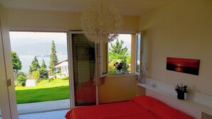 A bedroom with a red bedspread, a view of a garden and mountains through a sliding door, and a white ceiling light. - Africa apartment over Stresa with lake view (Stresa)