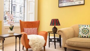 Una habitación con un ventanal grande, una silla con una manta de piel blanca, una lámpara sobre una mesita y un sofá con cojines estampados.