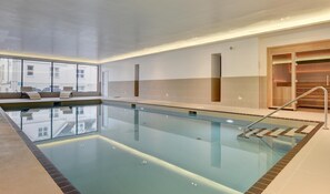 A modern indoor swimming pool with clear glass walls, a wooden sauna area, and a seating area with benches.
