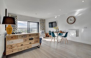 A modern living room with a wooden cabinet, a dining area with blue chairs, a large window, and a wall-mounted clock.