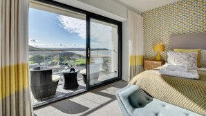 A hotel room with a large sliding glass door leading to a balcony, a bed with a patterned headboard, and a small side table with a lamp.
