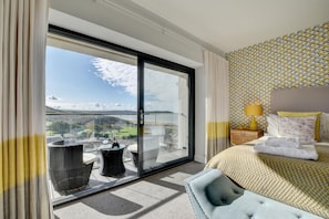 A hotel room with a large sliding glass door leading to a balcony, a bed with a patterned headboard, and a small side table with a lamp. - 3 Bed in Woolacombe (oc-5byron) (Woolacombe)