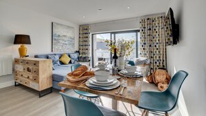 A modern living room with a sofa, a wooden sideboard, a dining table with chairs, and a television mounted on the wall.