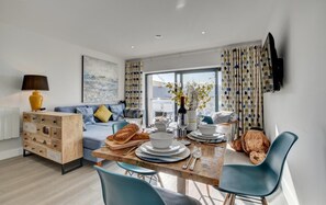 A modern living room with a sofa, a wooden sideboard, a dining table with chairs, and a television mounted on the wall.
