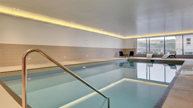 A modern indoor swimming pool with clear water, a metal ladder, and a seating area with chairs.