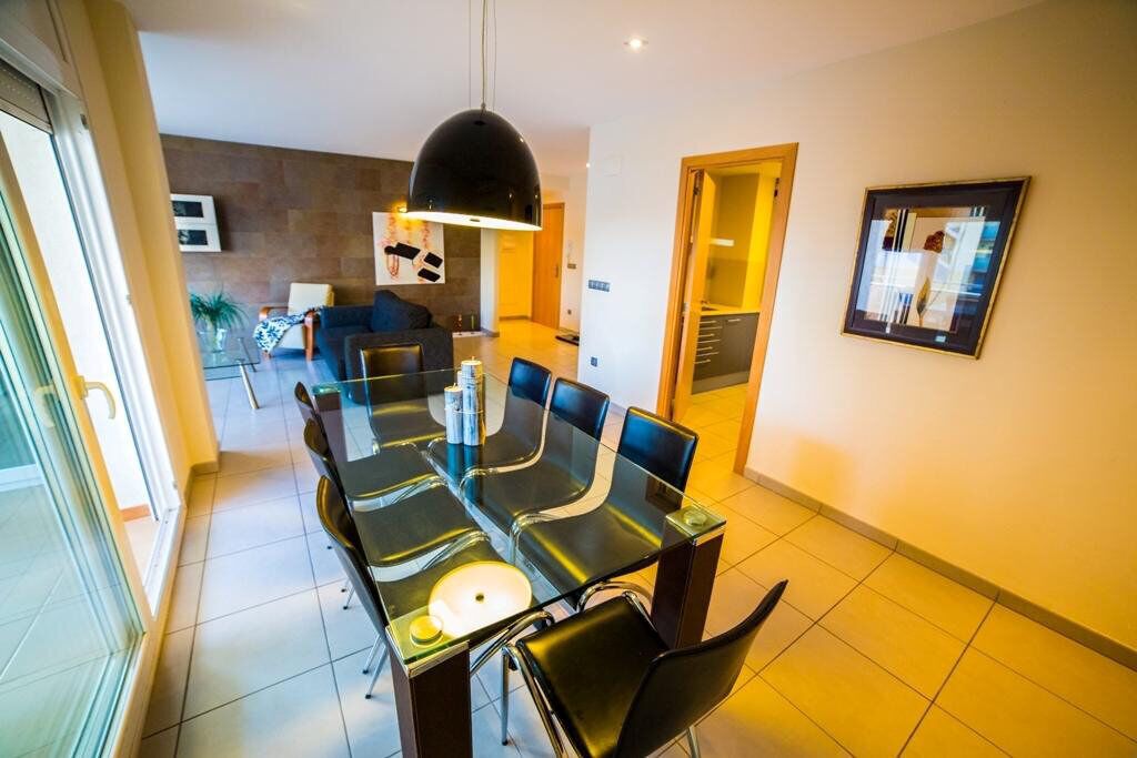 A modern dining area with a glass table and black chairs, a pendant light, and a framed picture on the wall.