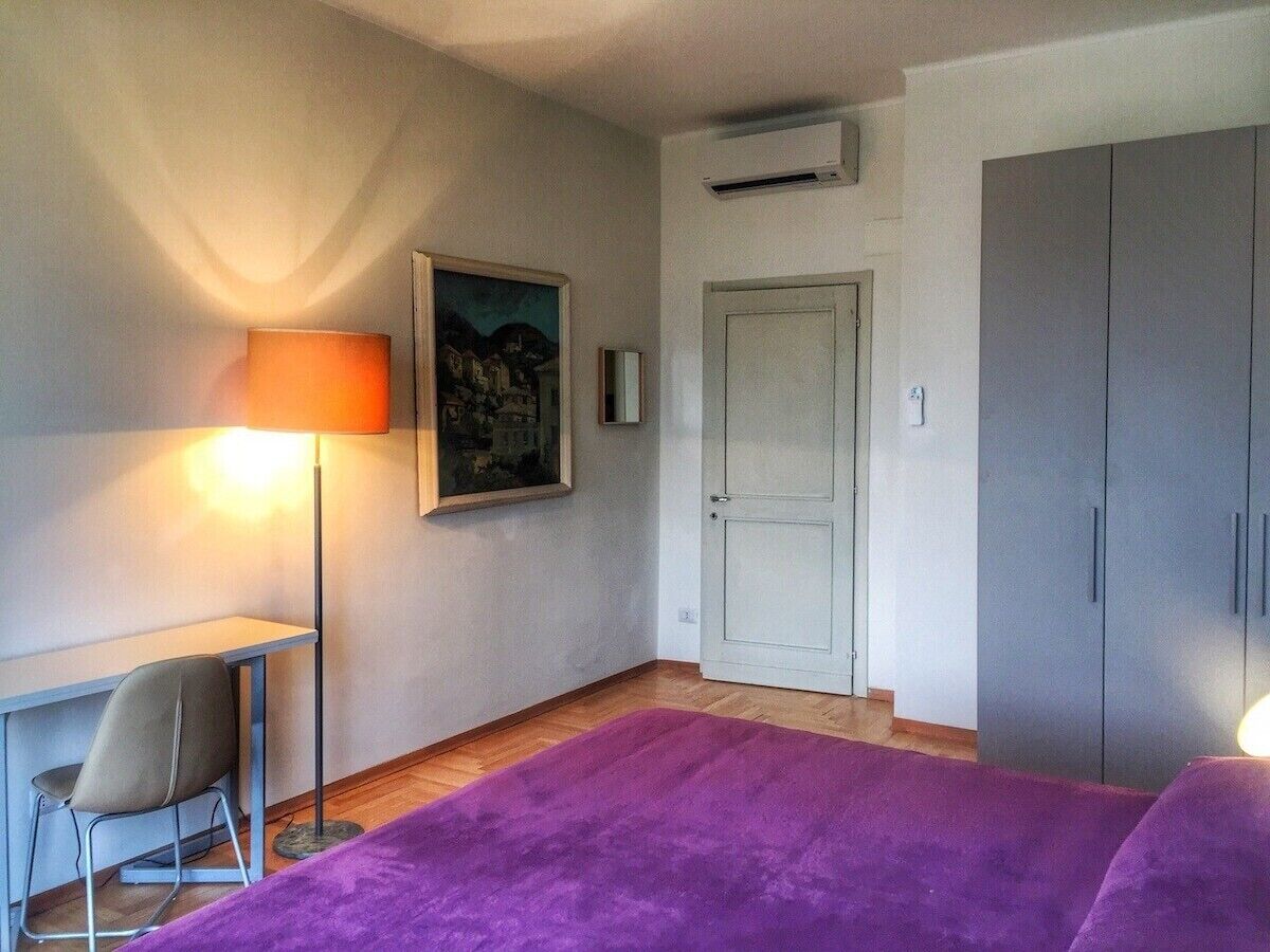 A room with a bed, a desk, a chair, a lamp, a painting, a wardrobe, and an air conditioning unit.