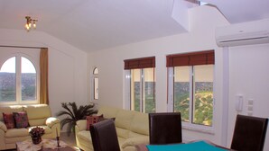 A living room with a sofa, a coffee table, and a desk with a chair. There are large windows with curtains, and an air conditioning unit is mounted on the wall. - Luxurious Private Villa with Private Pool and Sea Views- 6 min drive from beach (gouves)