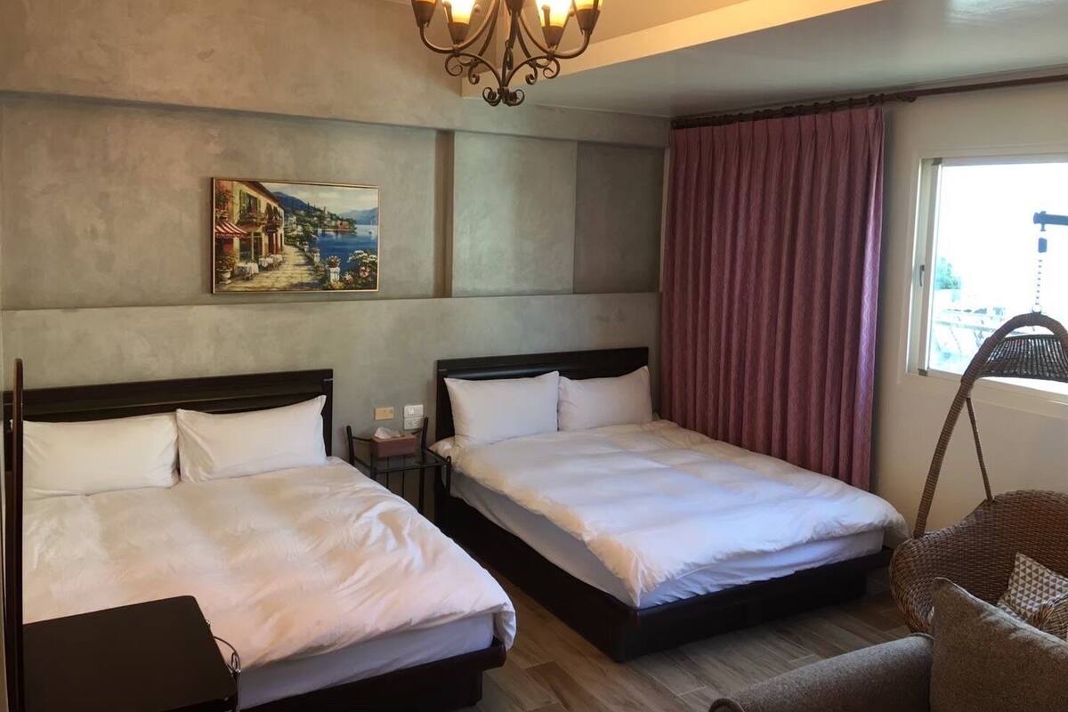 Habitación de hotel con dos camas, un cuadro en la pared, una lámpara de araña, una ventana con cortinas y una silla de mimbre.