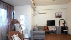 A living room with a grey sofa, a wicker chair with a stuffed toy, a TV, a small table with a floral tablecloth, and a desk with a computer and decorative items. - Beauty Home Homestay (Hualien City)