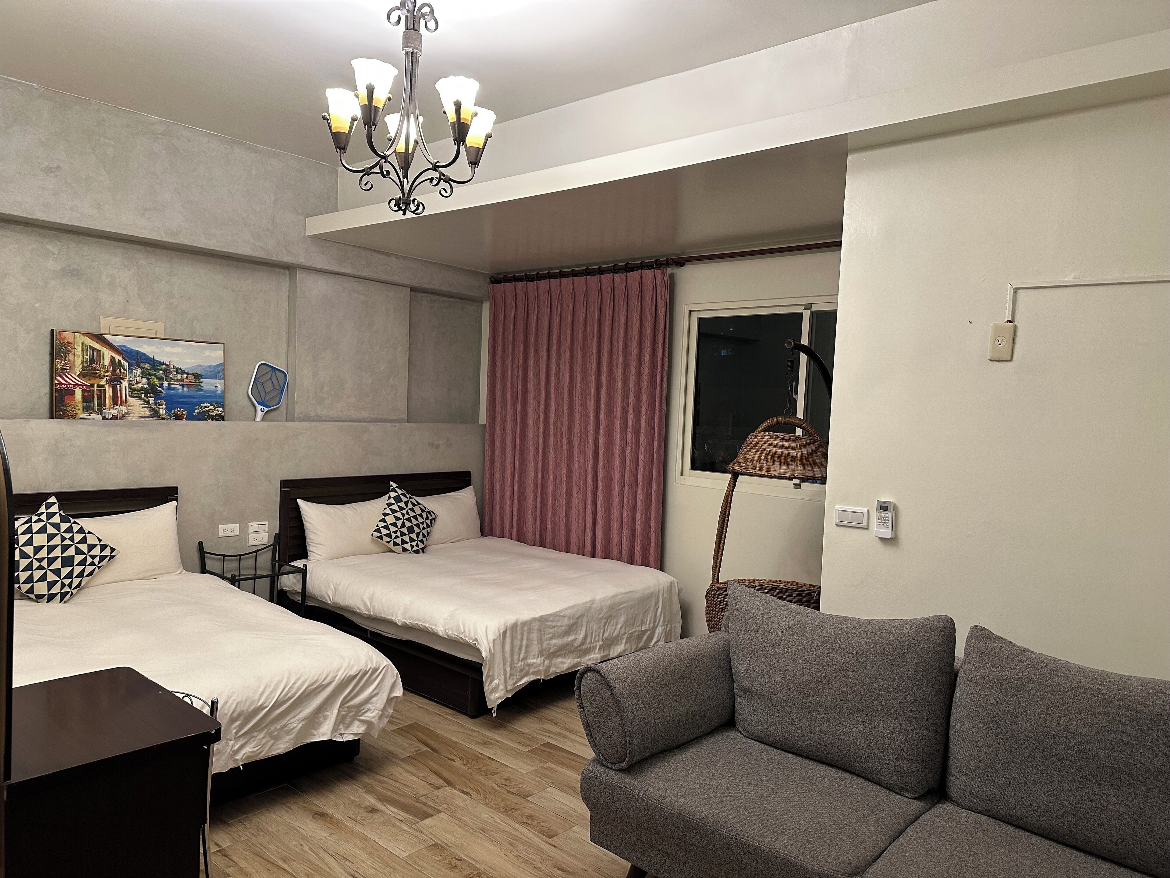 A hotel room with two beds, a sofa, a chandelier, a painting, and a window with curtains.