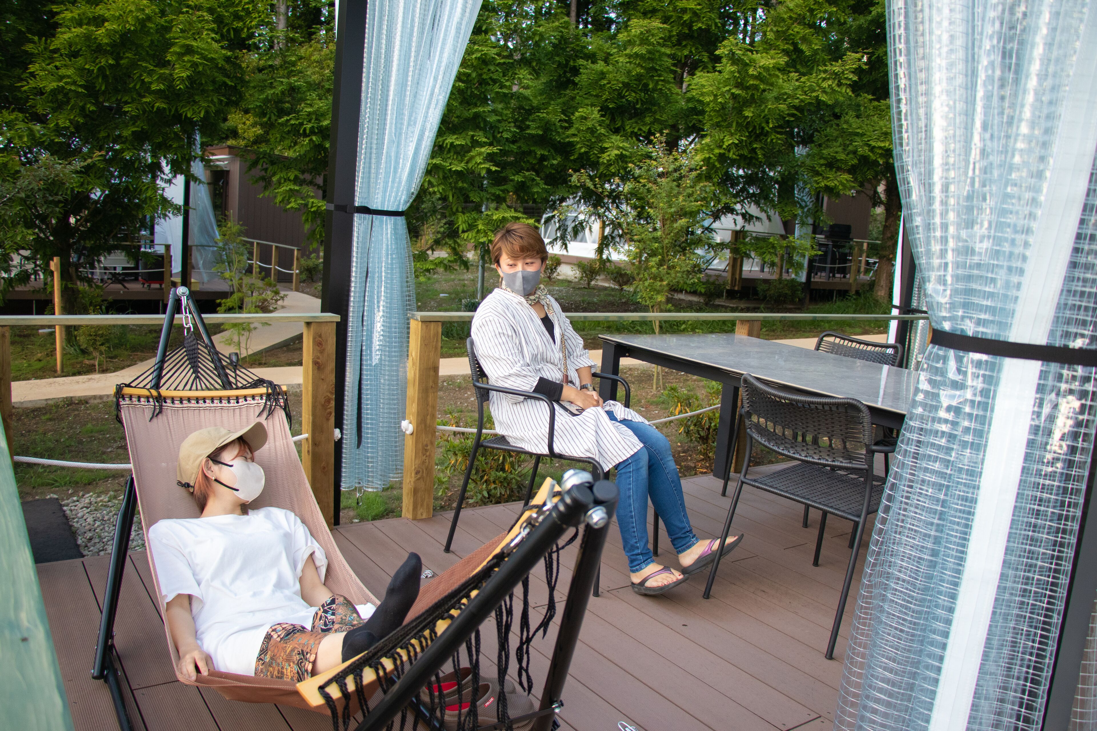 A wooden deck with outdoor seating, including a hammock and chairs, surrounded by greenery.